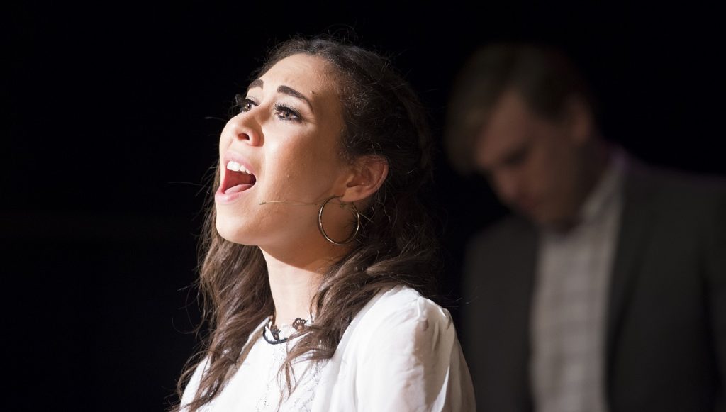 A musical theatre student sings, illuminated by a spotlight.