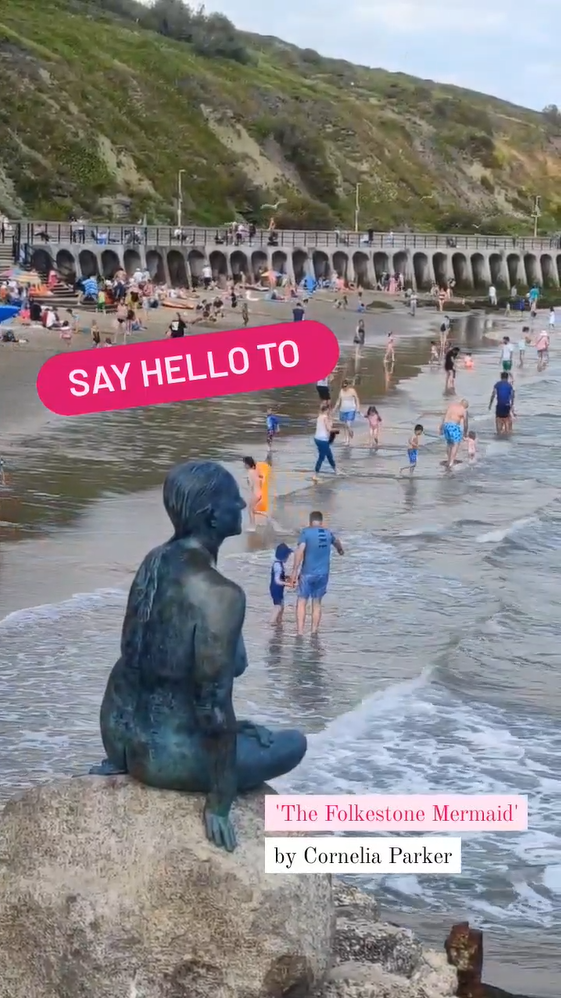 The Folkestone Mermaid by Cornelia Parker. A bronze sculpture on rocks beside a beach with families playing in the background.