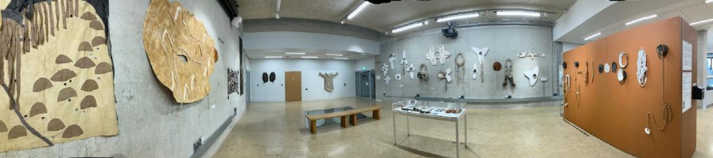 Wide angle view of the Daphne Oram Gallery, a large concrete-walled space, filled with artwork in neutral, natural tones. 