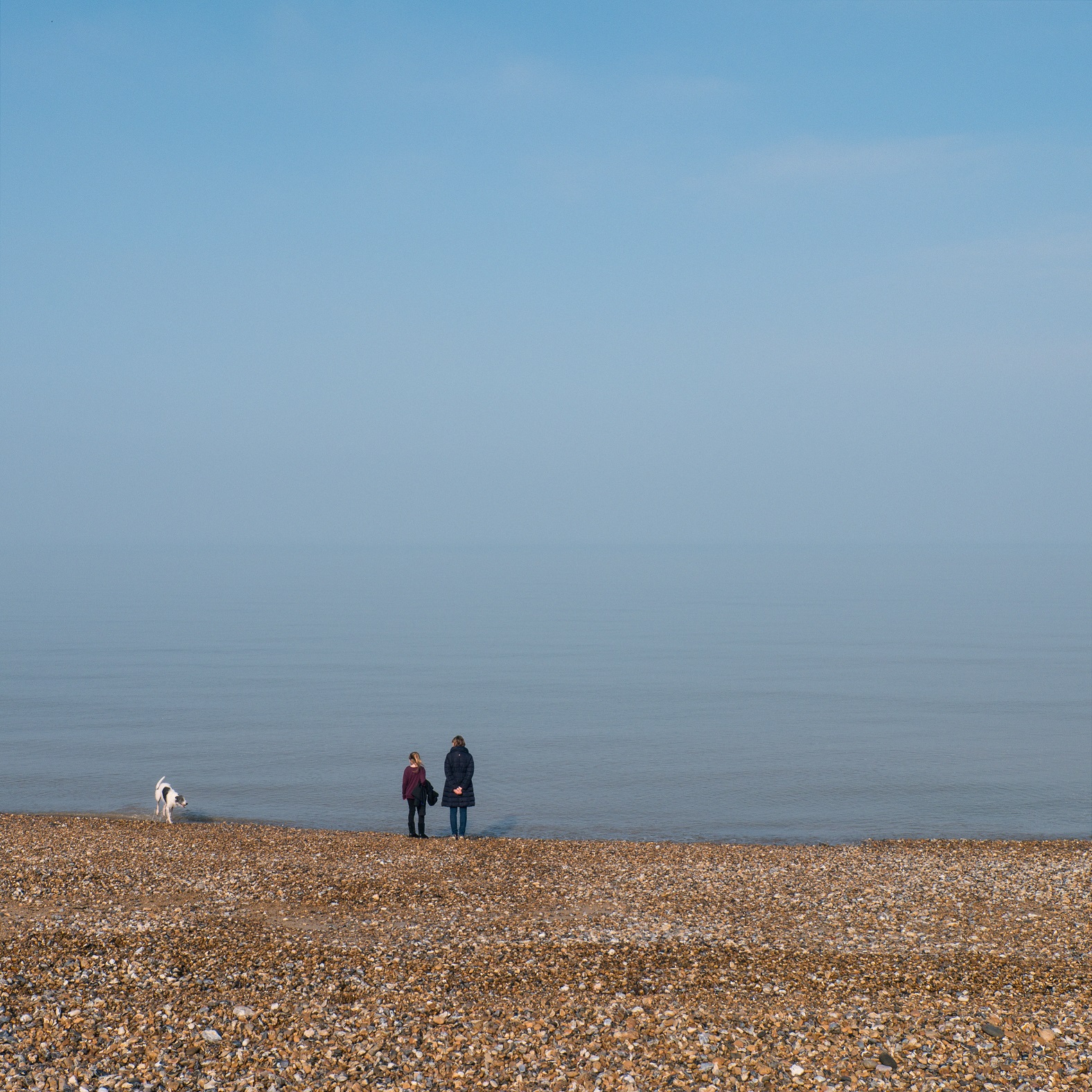 A colour photo of a stony beach, looking out across a still sea and hazy sky, with two figures and a dog at the water's edge.