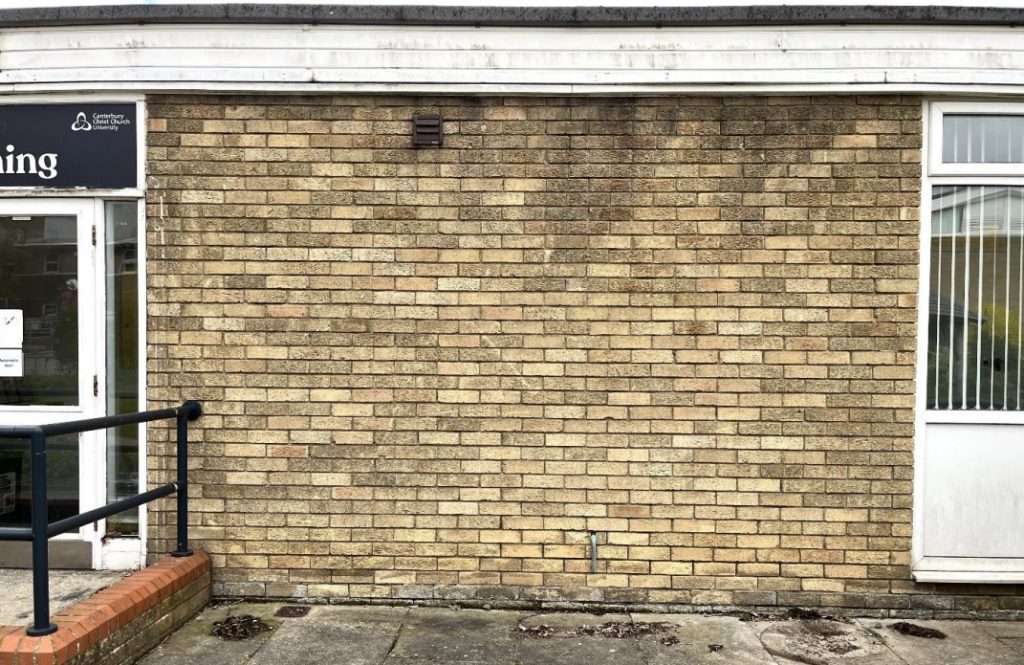 A single-storey, aged yellow brick wall on the CCCU campus, border by a door and railing to the left and a full height window to the right.