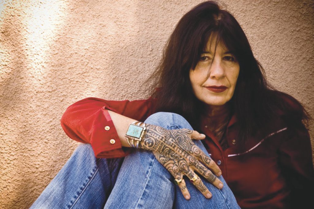 Smiling Joy Harjo in a red shirt and blue jeans, sitting in front of a light pink wall and showing off her whole right hand Marquesas Islands tattoo