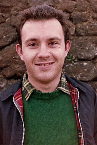 A photograph of a smiling Mark Ashton taken in 1986. He is standing against a brick wall. and wearing a checked shirt, green jumper and navy jacket.