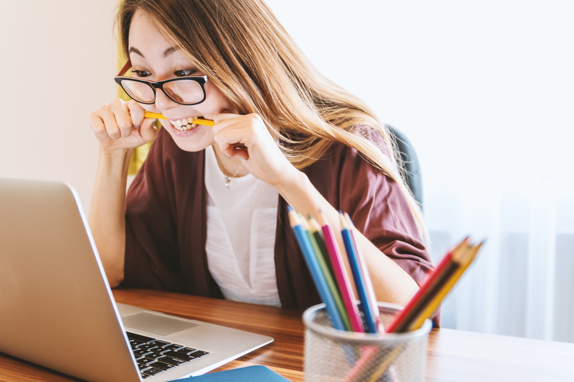 A young woman, with long hair and glasses, sitting at her desk. She is staring at her laptop screen while biting through a pencil