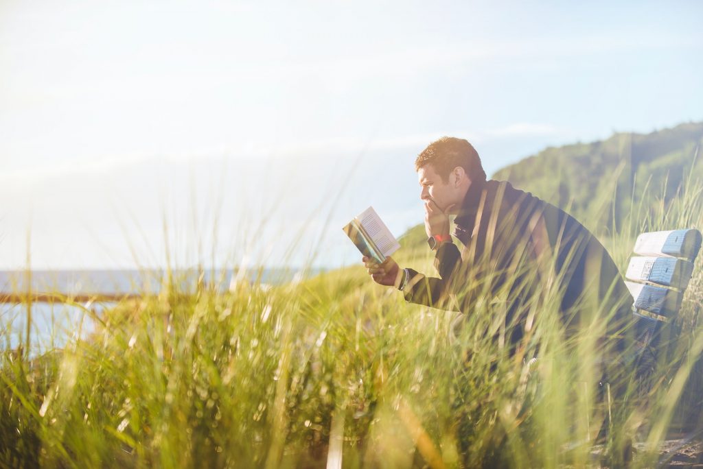 A man sitting on a bench, reading a book, by the coast.