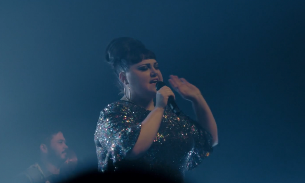 Beth Ditto live on stage in a sequin dress, singing into a microphone on a smoky stage