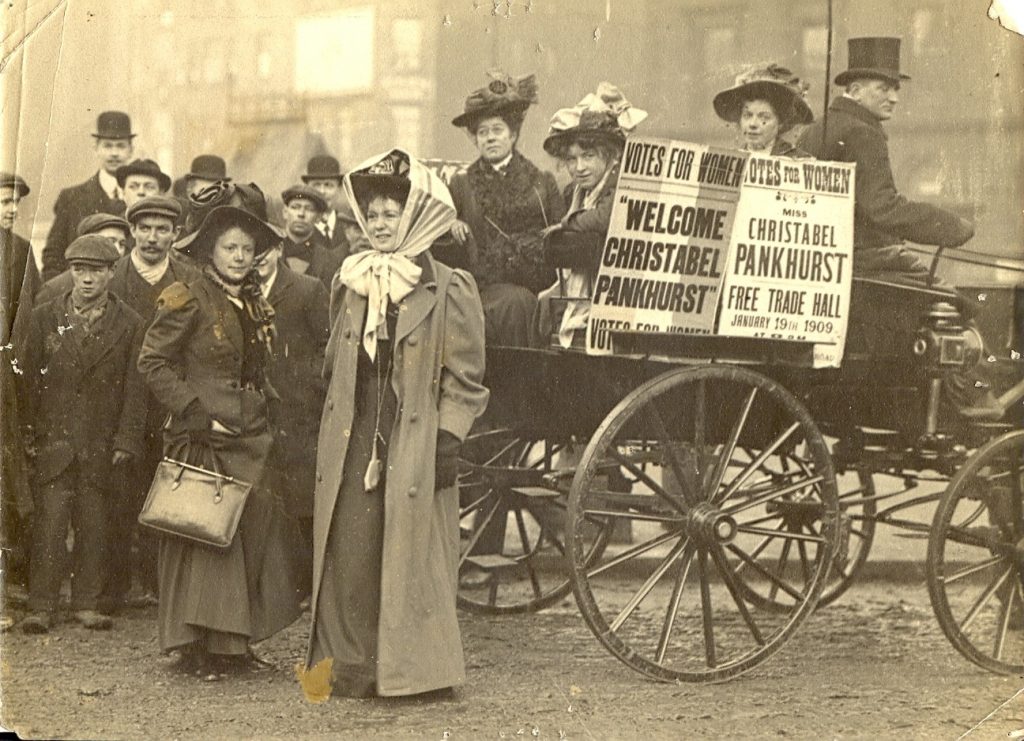 Men and women standing behind a drawn horse and coach. A women on the coach is holding two Votes for Women posters. 