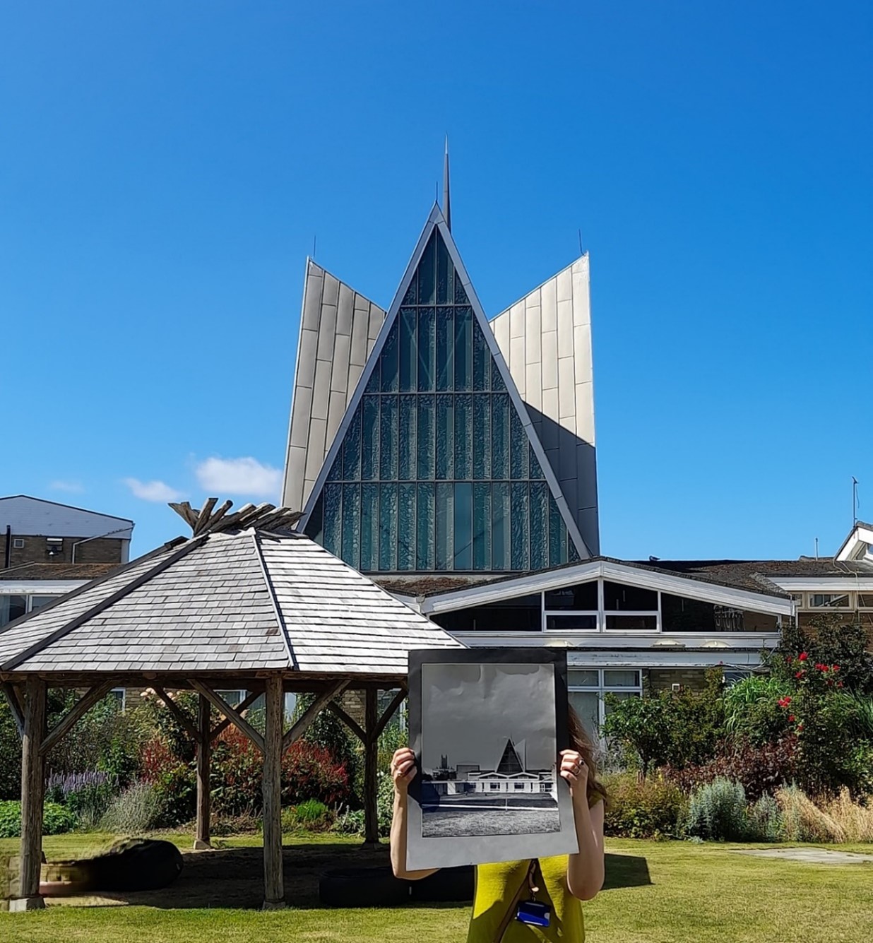 A figure holds up a black and white photo from the 60s covering their face, with the current day building on campus behind them in colour with a bright blue sky