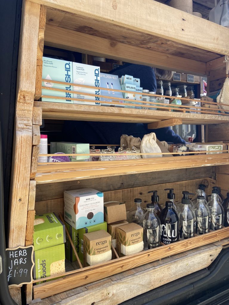 Photo of a range of bottles, packets and powders on wooden shelves for sale from the refillable van.