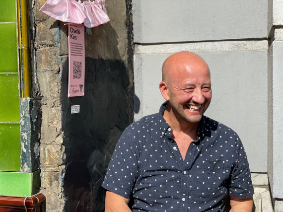 a man in a dark blue patterned short sleeve shirt sits smiling joyfully in the sunshine ouside an exhibit with his name, Charlie Kiss, on a plaque