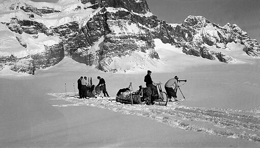Black-and-white photograph of Antarctic mountain scenery, with two sledges in the foreground. One man is bent forward using a survey instrument, whilst team member stands behind to record the data.