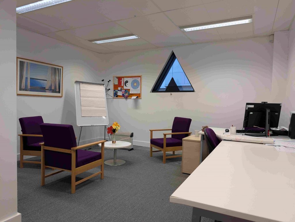 The inside of AHg.49, taken before it's renovations to become the Family Study Room. It is a bright room with white walls, three purple chairs and a desk with a PC.