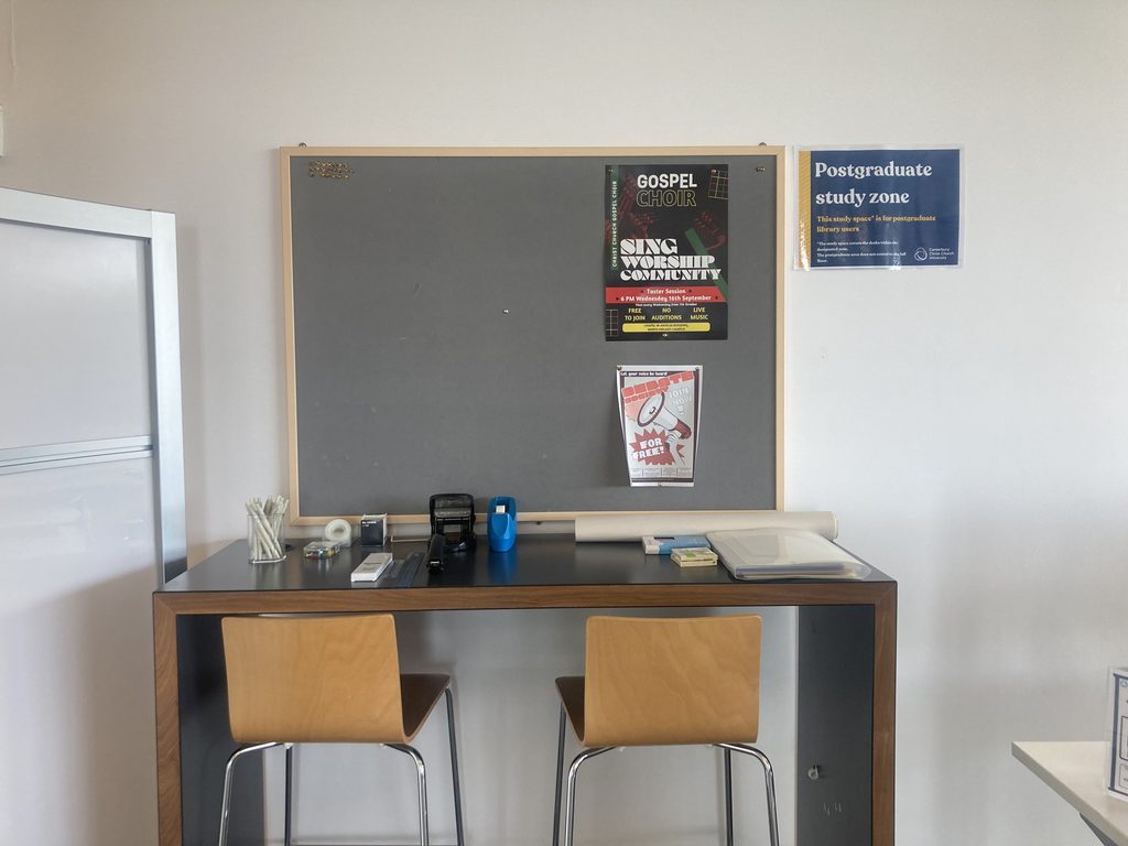 A desk with two stools and stationary, and a notice board with two posters detailing university events.
