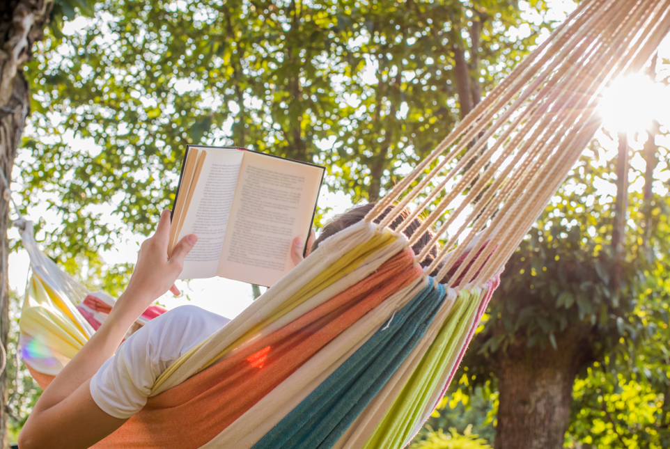 A photograph of a person reading a book on a hammock, surrounded by greenery and trees.