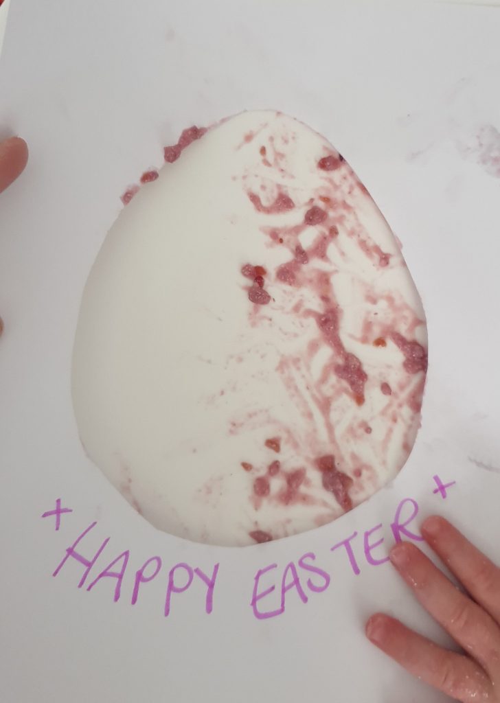 White Paper with an egg shape cut in middle held over edible paint so you can see the paint colour through the cut out. Text below read Happy Easter