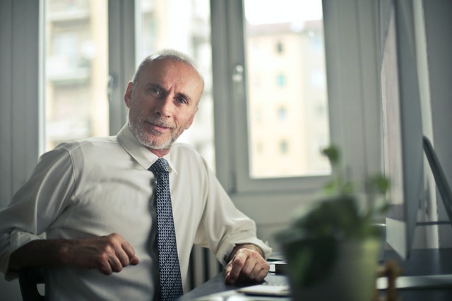 Man sitting at desk. There is a plant pot in the foreground out of focus. 